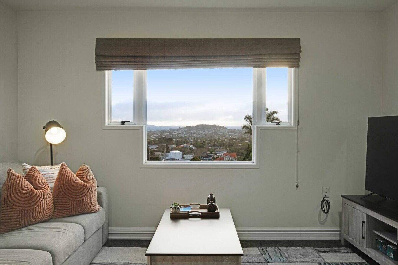 A living room with a sofa, a coffee table, a lamp, a TV, and a window with a view of a town.