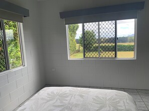 Una habitación con paredes de ladrillo blanco, una cama con colcha floral y una ventana con vistas a zonas verdes y colinas.