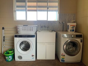 A laundry room with two washing machines, a drying machine, a metal drying rack, a bucket, and a window with a view.