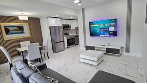 Una sala de estar moderna con sofá, mesa de comedor y sillas, una cocina con electrodomésticos, y un televisor montado que muestra servicios de transmisión.