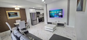 Ein modernes Wohnzimmer mit einer Couch, einem Esstisch und Stühlen, einer Küche mit Geräten und einem montierten Fernseher, der Streaming-Dienste anzeigt.
