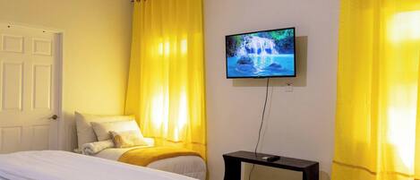 A hotel room with a bed, a TV mounted on the wall, and a window with yellow curtains.