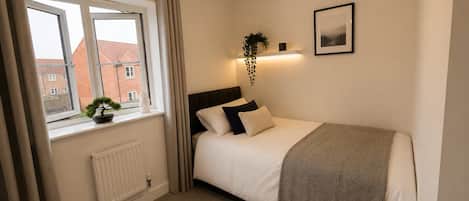 A bedroom with a bed, a window, a radiator, a plant, a picture on the wall, and a light fixture.