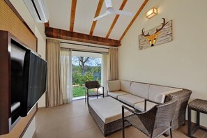 A living room with a TV, sofa, wicker chairs, a ceiling fan, and a view through a glass door. - The Fern Sattva Resort Polo Forest (Vijayanagar)
