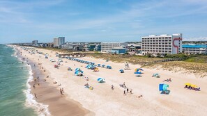 A beach with umbrellas and people, a pier, and buildings in the background. - Luxury Ocean View Home w Pool Access, Rooftop, and Pool Table! (Carolina Beach)