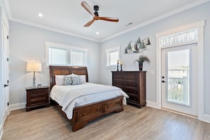 A bedroom with a wooden bed, a ceiling fan, a nightstand with a lamp, a dresser, and a door with a view. - Luxury Ocean View Home w Pool Access, Rooftop, and Pool Table! (Carolina Beach)