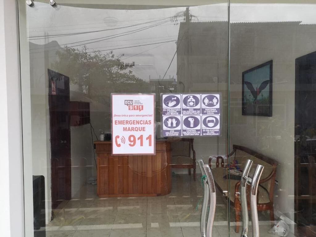 A reception desk with a sign indicating emergency contact information and a glass door reflecting a building and a bird.
