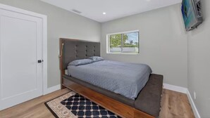 Un dormitorio con cama, ventana, puerta y televisor montado en la pared.