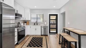 Una cocina moderna con muebles blancos, electrodomésticos de acero inoxidable, una alfombra negra y beige, y un comedor con banco y mesa.