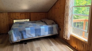 Et soveværelse med træloft, et vindue med udsigt til grøn natur og en seng med et mønstret sengetæppe.