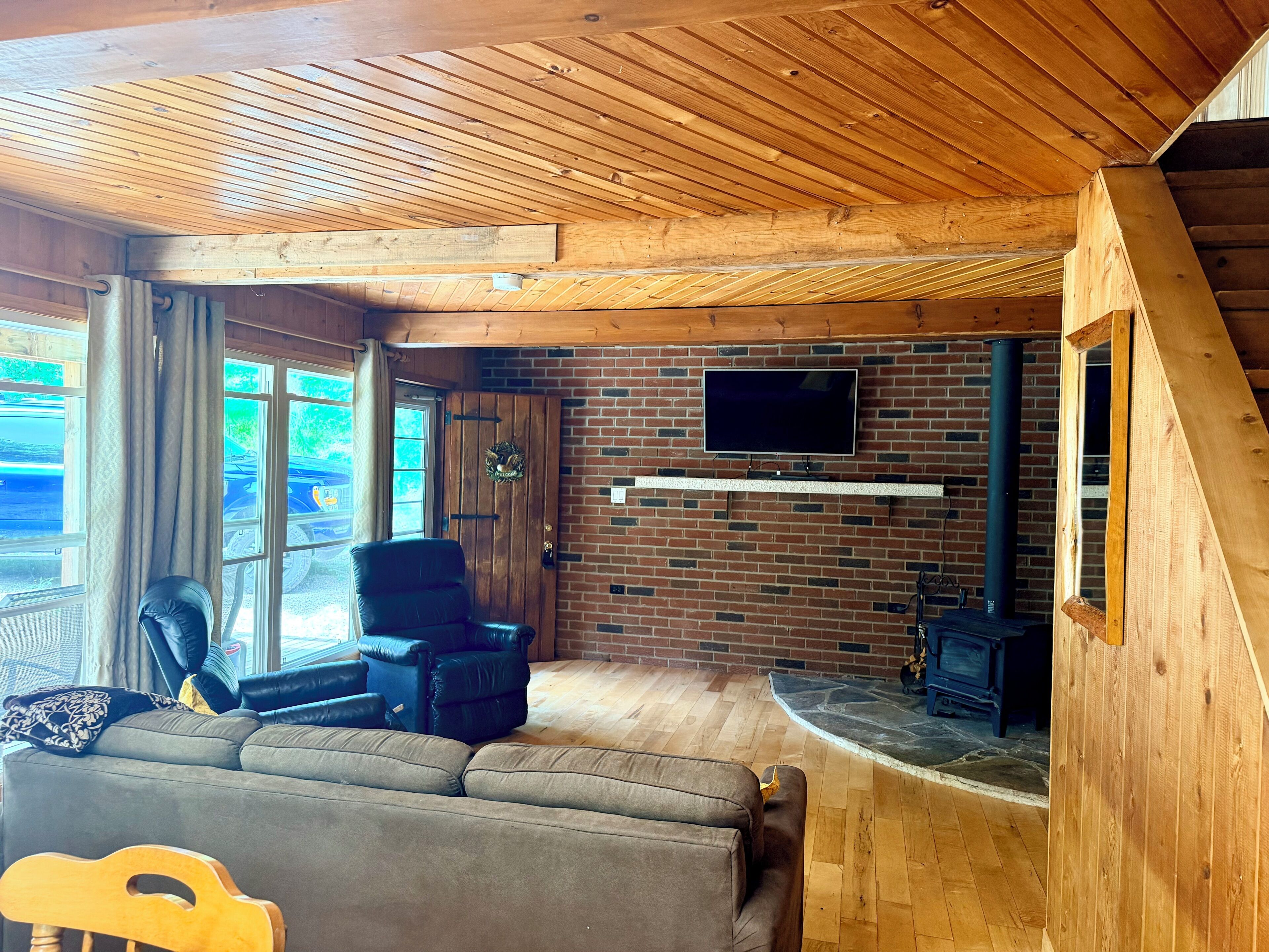 Een gezellige woonkamer met houten plafond, een schouw van baksteen, een aan de muur bevestigde televisie en een leren bank.