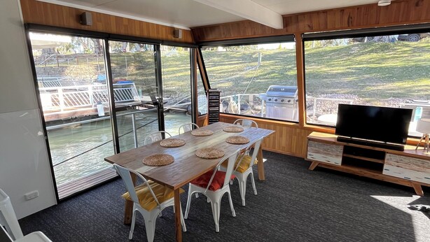 A dining area with a long table, white chairs, and a television on a wooden stand. - 3-bedroom house boat in beautiful Swan Reach with beautiful Murray River views. (Punyelroo)