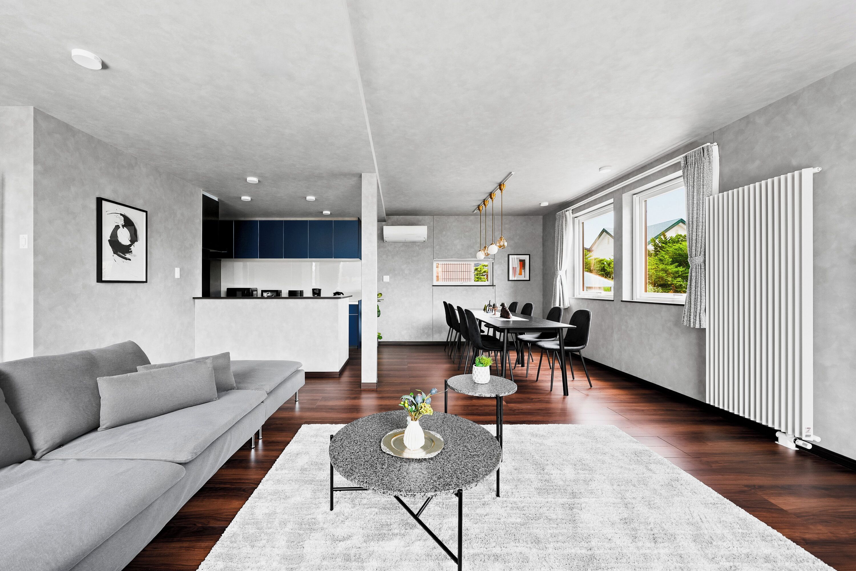 Ein modernes Wohnzimmer mit einem grauen Sofa, einem runden Couchtisch mit einer Vase, einem Essbereich mit Stühlen und einer Küche im Hintergrund.