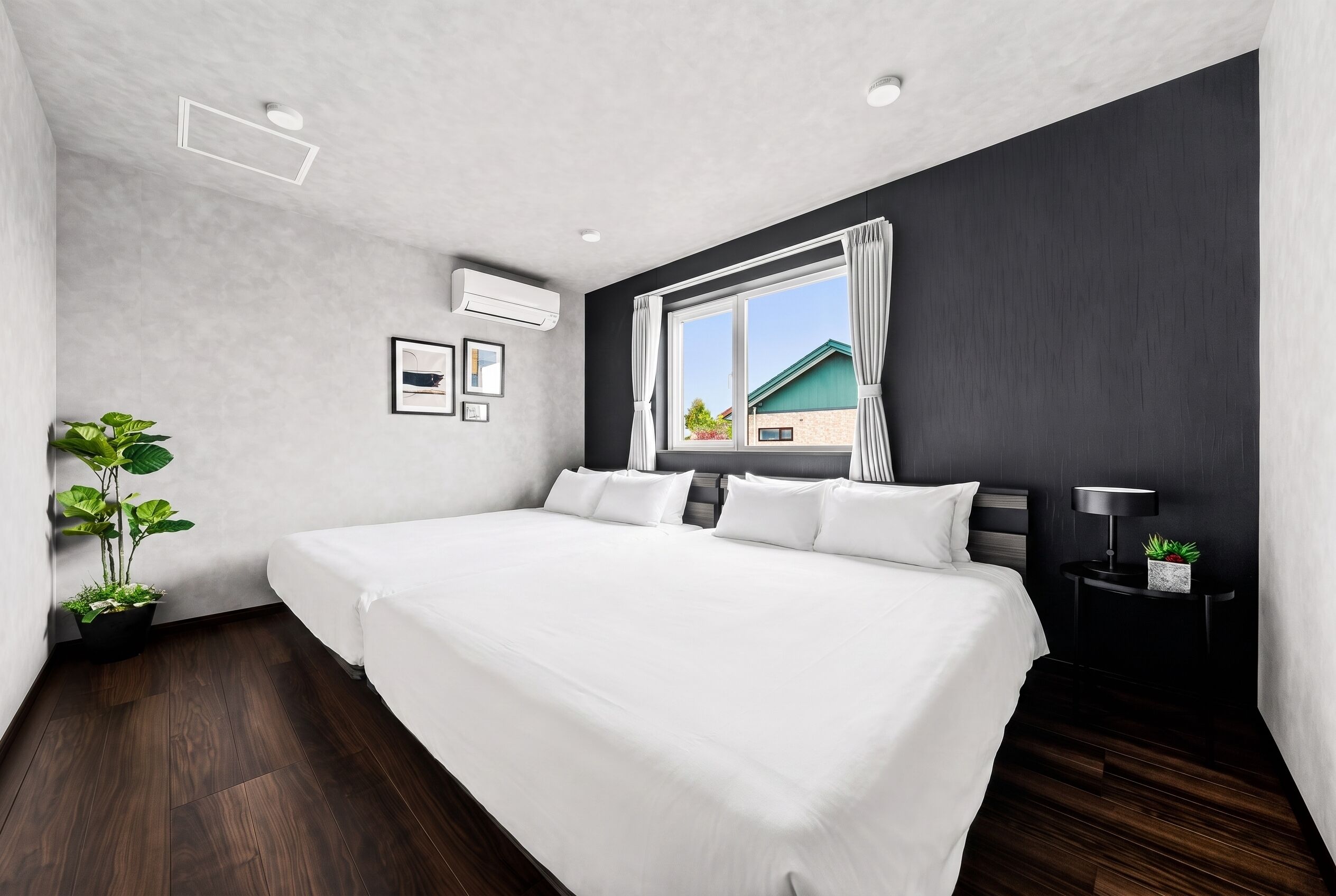 A modern bedroom with a large bed, a potted plant, and a view of a house through a window.