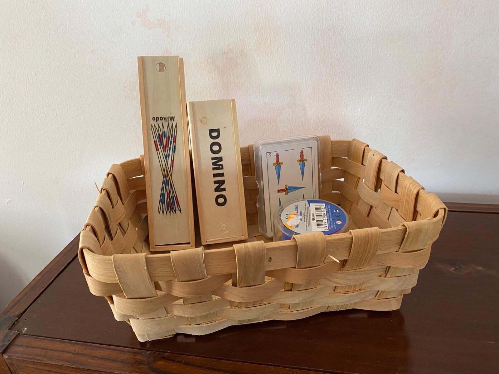 A woven basket containing a wooden block with 'DOMINO' engraved, a small plastic container with a label, and a card with a game illustration.