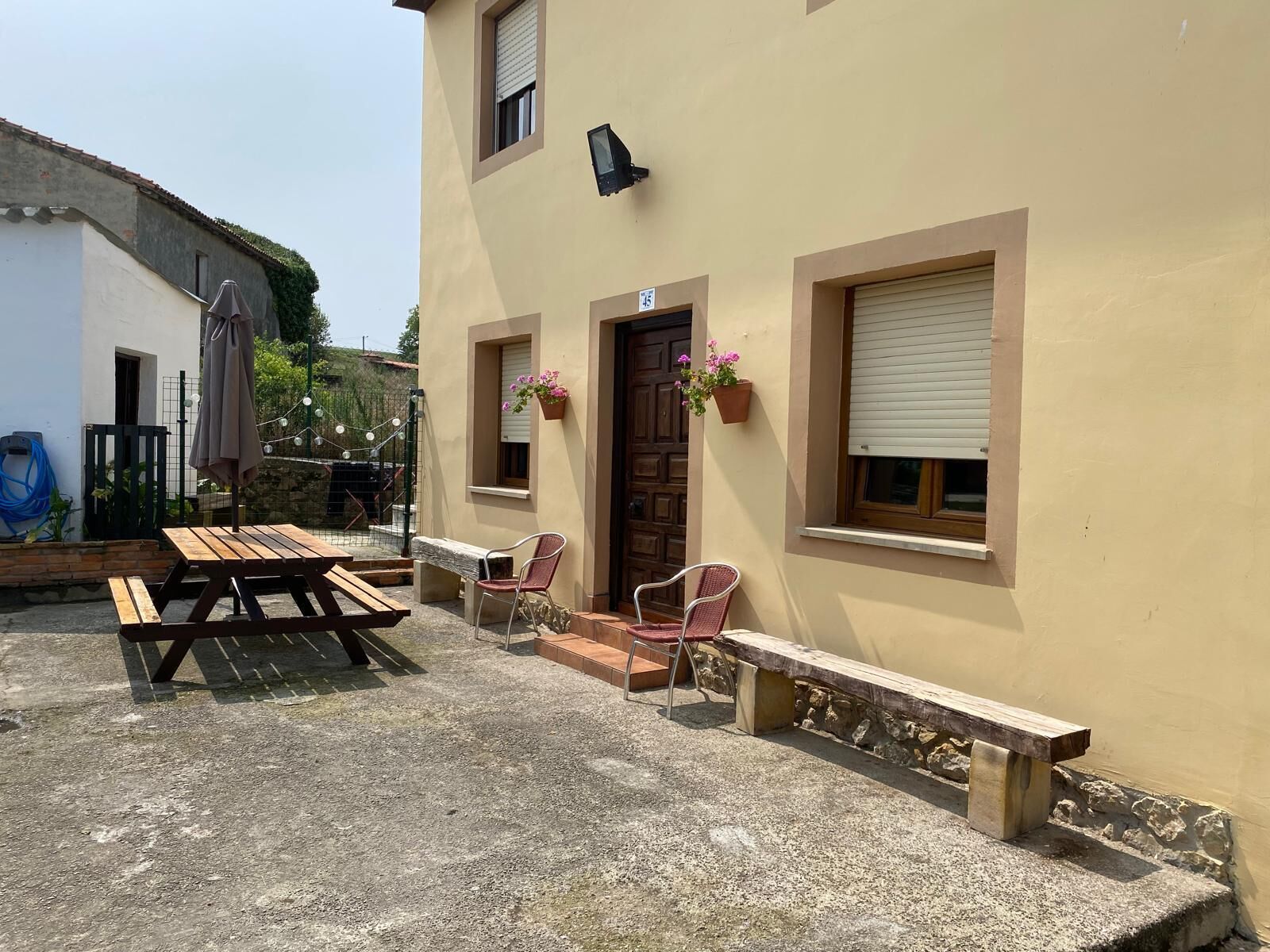 A building with a patio area featuring a wooden table and chairs, a door with a window, and a small flower pot on the wall.