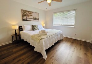 A bedroom with a bed, a ceiling fan, a window with blinds, a nightstand with a lamp, and a painting on the wall. - Marblehead Executive Retreat (Jackson)