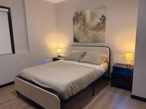 Une chambre à coucher comprenant un lit, deux tables de chevet, un tableau abstrait accroché au mur et une fenêtre avec des stores.