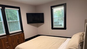 Quarto com cama, televisão fixada na parede e duas janelas com persianas.