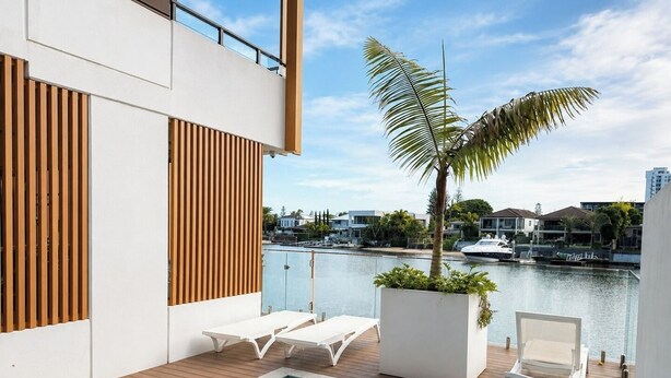Un área de piscina moderna al aire libre con cubierta de madera, sillones blancos y vista a un puerto deportivo con barcos.