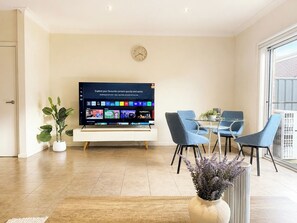 A modern living room with a flat-screen TV, a dining area with blue chairs, a potted plant, and a large window.