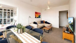 Ein modernes Wohnzimmer mit einem Glastisch, Sofas, einem Couchtisch, Deckenventilatoren, einem Bücherregal und einem Gemälde.