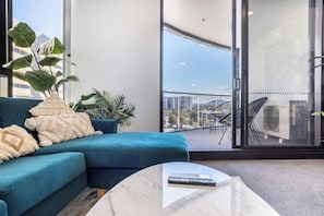 A modern living room with a blue sofa, a coffee table, and large windows offering a city view. - Elegant 3BR Retreat with Pool, Parking and Balcony (Phillip)