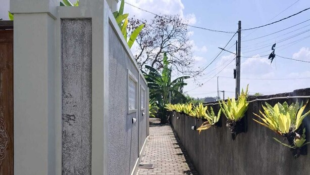 A narrow brick pathway flanked by a concrete wall with green plants, leading to a building with a tiled roof. - Cozzy Villa Sakangi Mas (Tabanan)