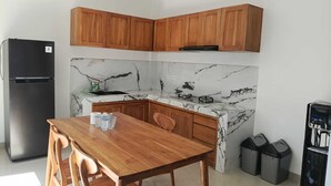 A compact kitchen with wooden cabinets, a marble backsplash, a black refrigerator, a dining table with chairs, and black trash bins. - Cozzy Villa Sakangi Mas (Tabanan)