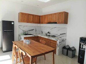Eine kleine Küche mit Holzschränken, einer Marmor-Spritzschutzwand, einem schwarzen Kühlschrank, einem Esstisch mit Stühlen und schwarzen Mülleimern.