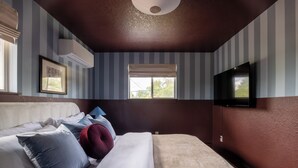 Una habitación de hotel con cama, televisor, ventana con persianas y equipo de aire acondicionado.