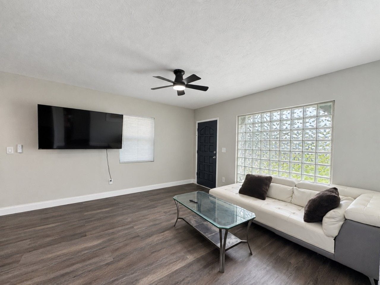 A living room with a flat-screen TV, a glass coffee table, a ceiling fan, a sofa with cushions, and a window with a decorative screen.