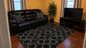 Una sala de estar con un sofá de cuero negro, dos almohadas a cuadros blanco y negro, un tapete con estampado hexagonal en negro, una planta en maceta, un televisor de pantalla plana sobre un soporte negro y un ventilador de techo con luces.