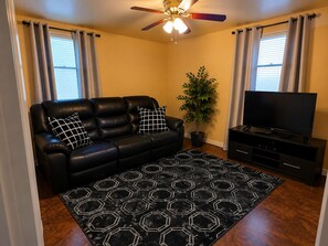 Ein Wohnzimmer mit einer schwarzen Ledersofa, zwei schwarz-weiß karierten Kissen, einem schwarzen, hexagonal gemusterten Teppich, einer Zimmerpflanze, einem Flachbildfernseher auf einem schwarzen Standfuß und einem Deckenventilator mit Beleuchtung.