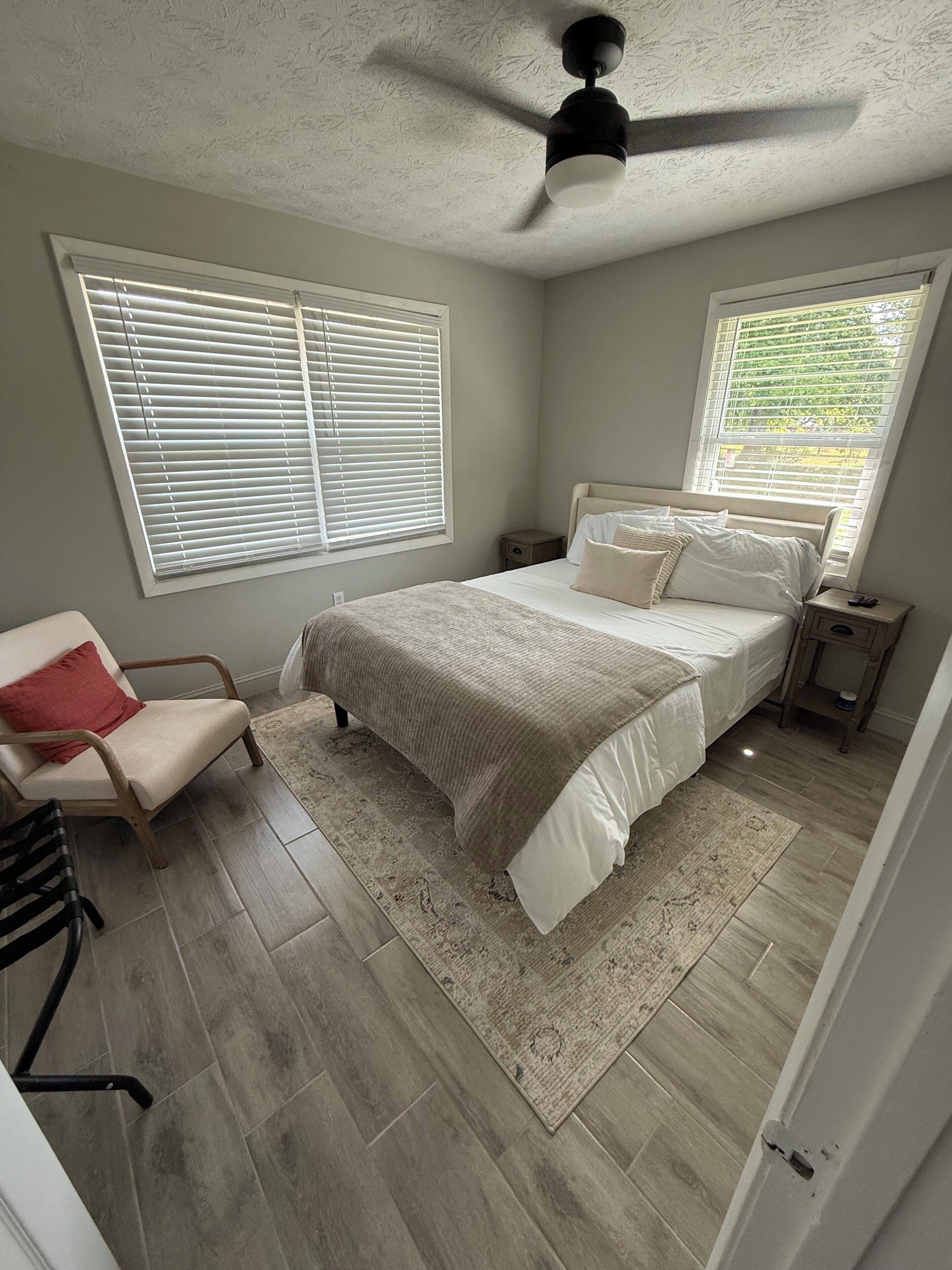 A bedroom with a bed, a chair, a nightstand, a ceiling fan, and large windows.