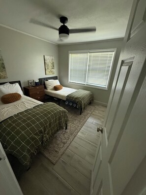 A bedroom with two beds, a ceiling fan, a window with blinds, and a door.