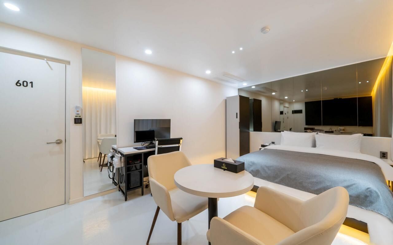A modern hotel room with a large bed, a desk with a computer, a chair, and a small table.