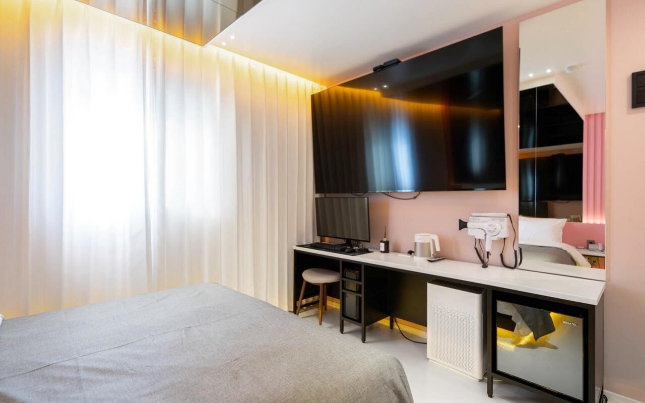 A modern hotel room with a bed, a desk with a computer, a mirror, and a television.