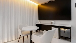 A modern room with a flat-screen TV mounted on the wall, a round white table, two white chairs, and a large window with vertical blinds. - Lizena HOTEL (Asan)