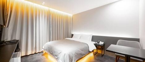 A modern hotel room with a bed, bedside table, chair, and a desk with a computer.