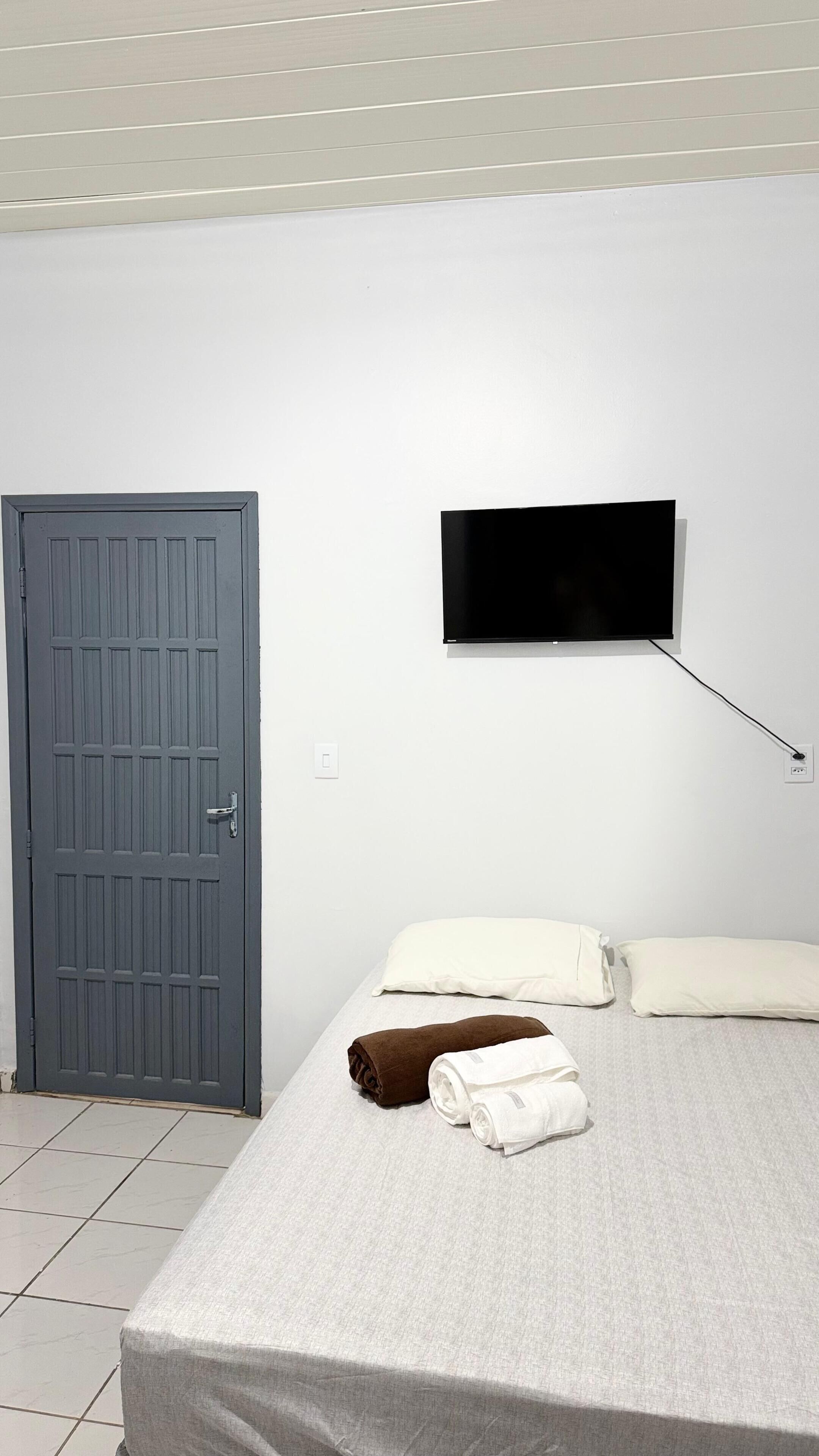 Une pièce avec une porte grise, un lit avec des serviettes blanches et brunes, un téléviseur à écran plat fixé au mur et un plafond blanc.