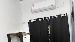Una habitació amb cortina negra, escriptori de fusta amb calaixos, nevera Philco negra i aire condicionat fixat a la paret.