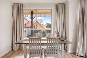 Un comedor con una mesa y sillas, una ventana con cortinas, y vistas a casas desde afuera.
