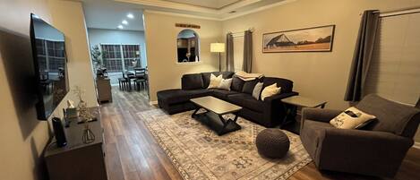 A modern living room with a sectional sofa, a coffee table, and a TV. There is a dining area in the background with a table and chairs.