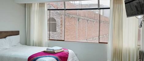 A hotel room with a bed, a chair, a television, and a window with a view of buildings.