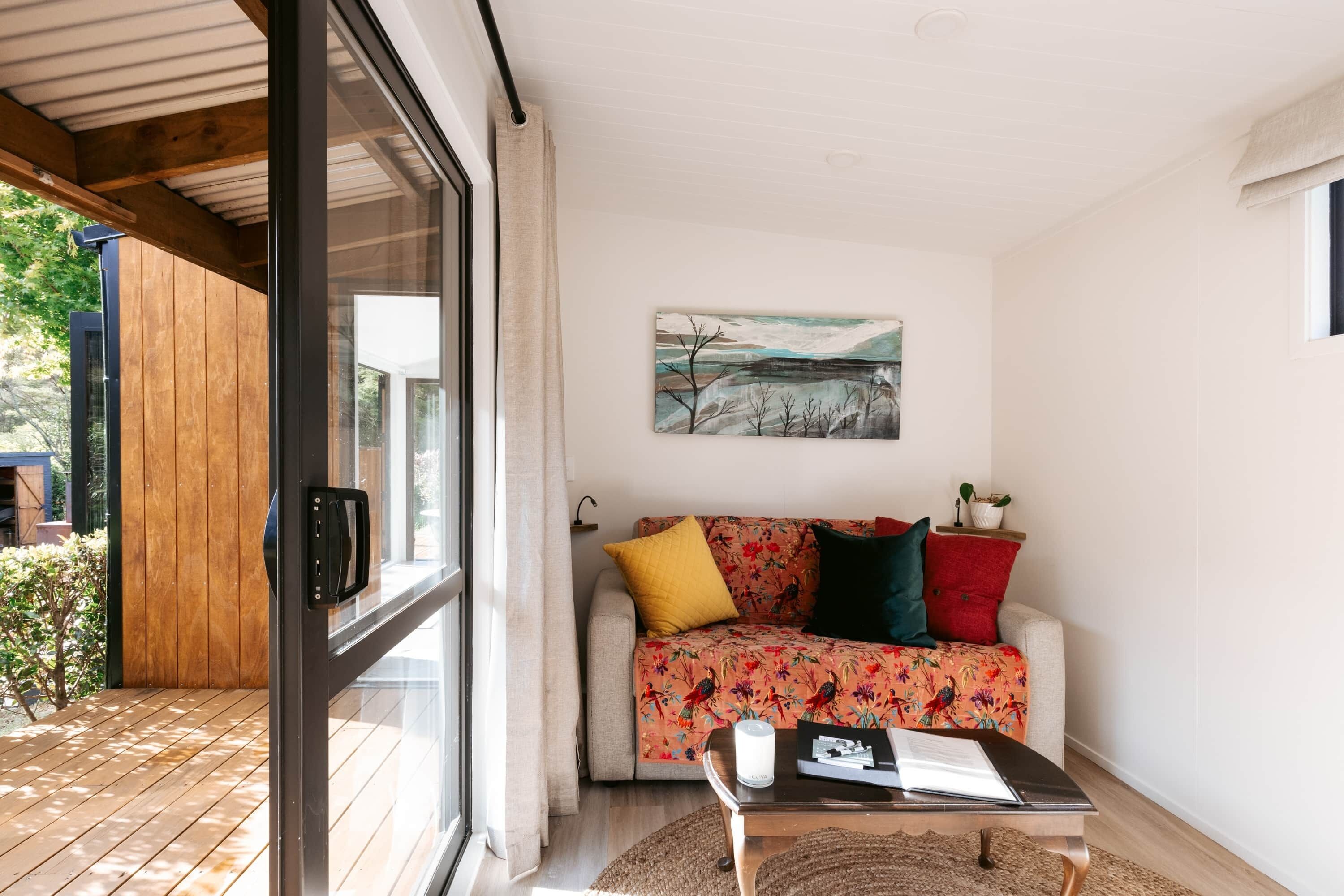 A modern living room with a wooden floor, a glass door, a sofa with colorful cushions, a painting on the wall, and a small table with a candle.