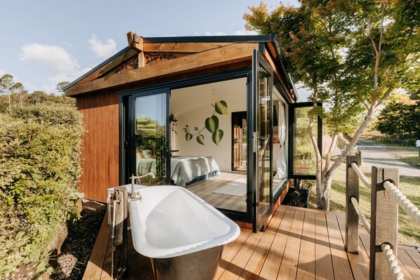 A modern, wooden cabin with a glass door leading to a bedroom, a wooden deck, and a large bathtub.