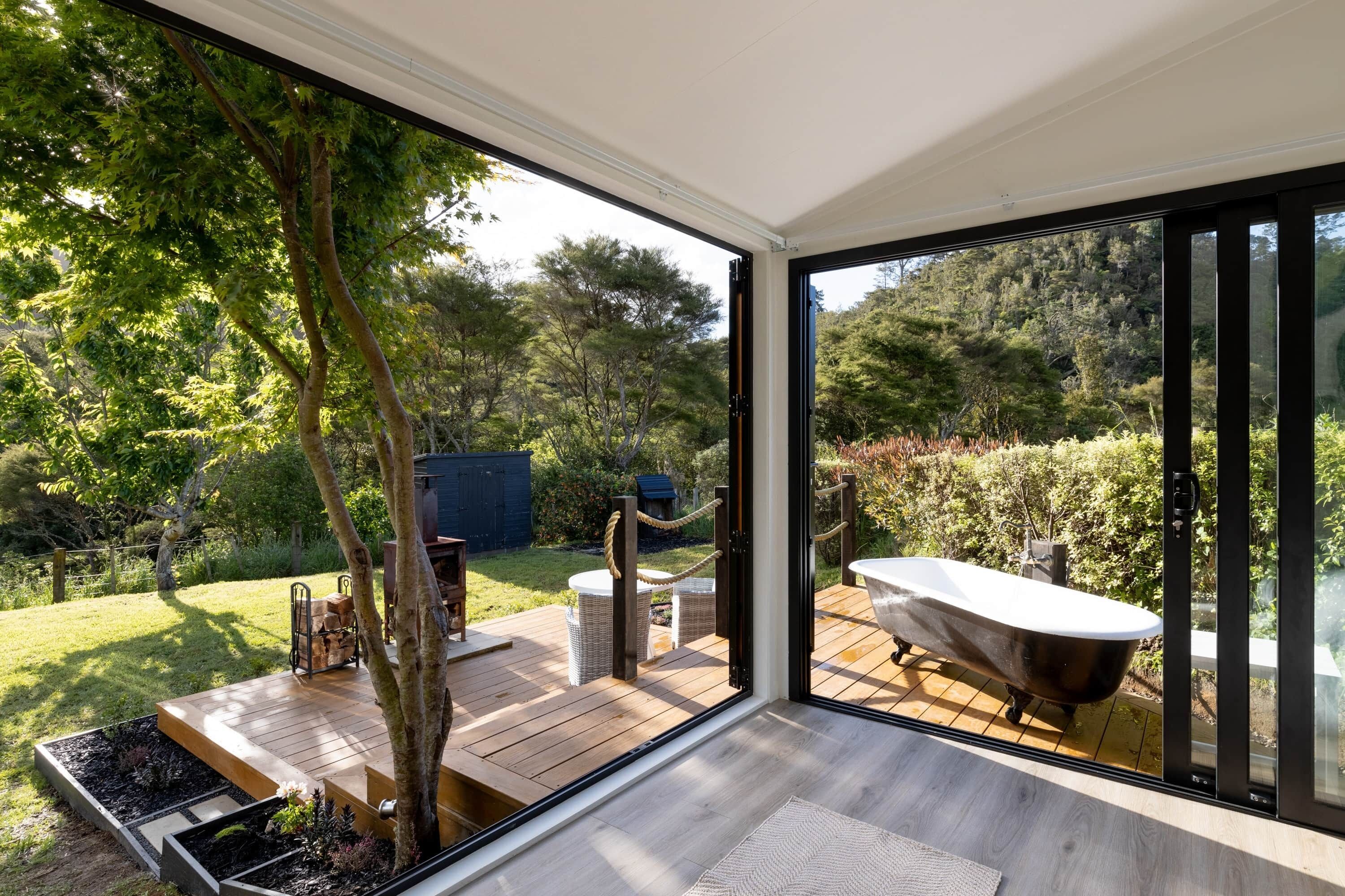 A modern bathroom with a freestanding bathtub, a wooden deck, and a glass door leading to a lush garden.