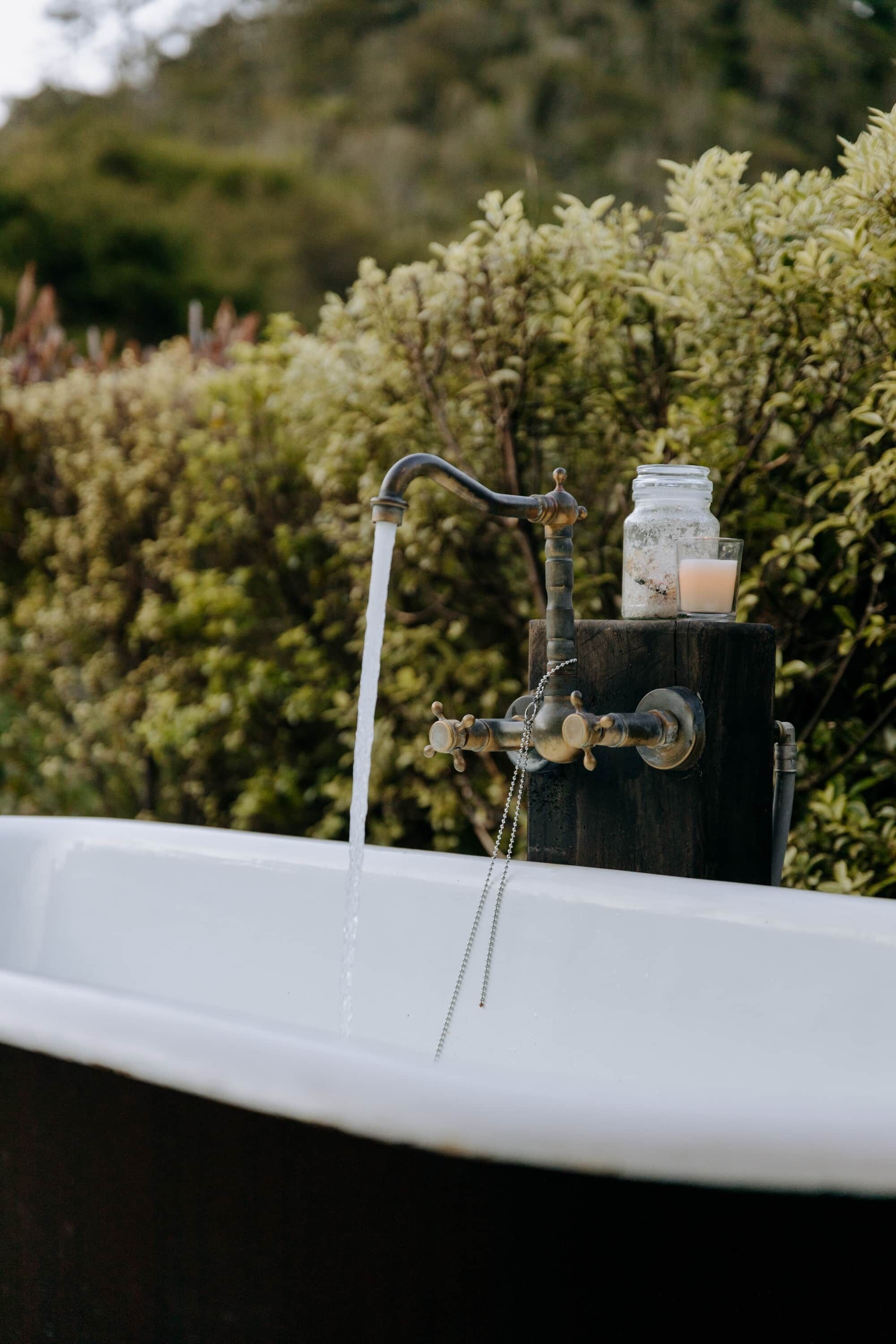 A bathtub with running water, a wooden stand with a jar and a candle, and a hedge in the background.