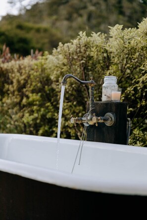 A bathtub with running water, a wooden stand with a jar and a candle, and a hedge in the background.